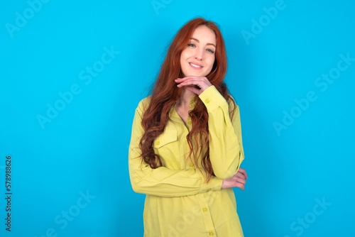 Optimistic young woman wearing green sweater over blue background keeps hands partly crossed and hand under chin, looks at camera with pleasure.