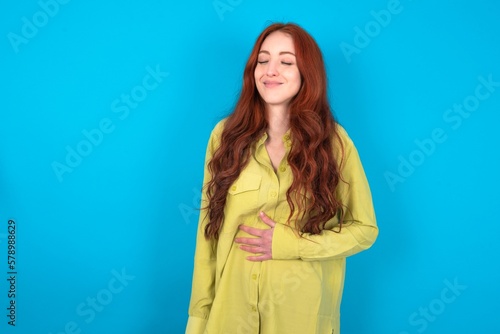 young woman wearing green sweater over blue background touches tummy, smiles gently, eating and satisfaction concept.
