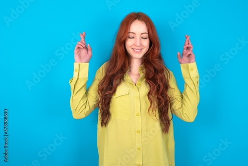 Konstfotografi young woman wearing green sweater over blue background has big hope, crosses fingers, believes in good fortune, smiles broadly
