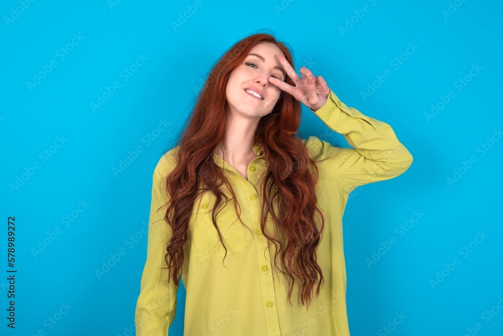 young woman wearing green sweater over blue background Doing peace symbol with fingers over face, smiling cheerful showing victory young woman wearing green sweater over blue background Doing peace symbol with fingers over face, smiling cheerful showing victory