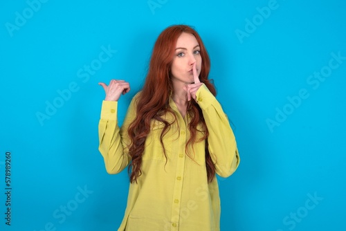 young woman wearing green sweater over blue background asking to be quiet with finger on lips pointing with hand to the side. Silence and secret concept.