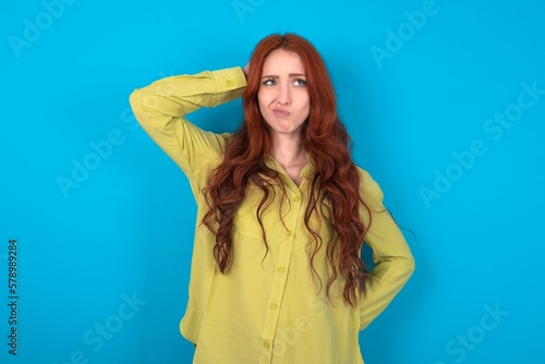 young woman wearing green sweater over blue background confuse and wonder about question. Uncertain with doubt, thinking with hand on head. Pensive concept.