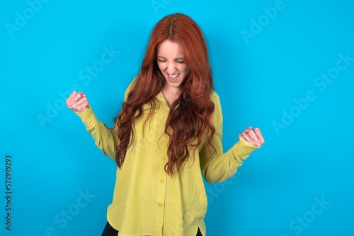 young woman wearing green sweater over blue background very happy and excited doing winner gesture with arms raised, smiling and screaming for success. Celebration concept.