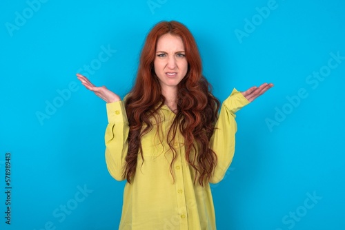 Indignant young woman wearing green sweater over blue background gestures in bewilderment, frowns face with dissatisfaction.