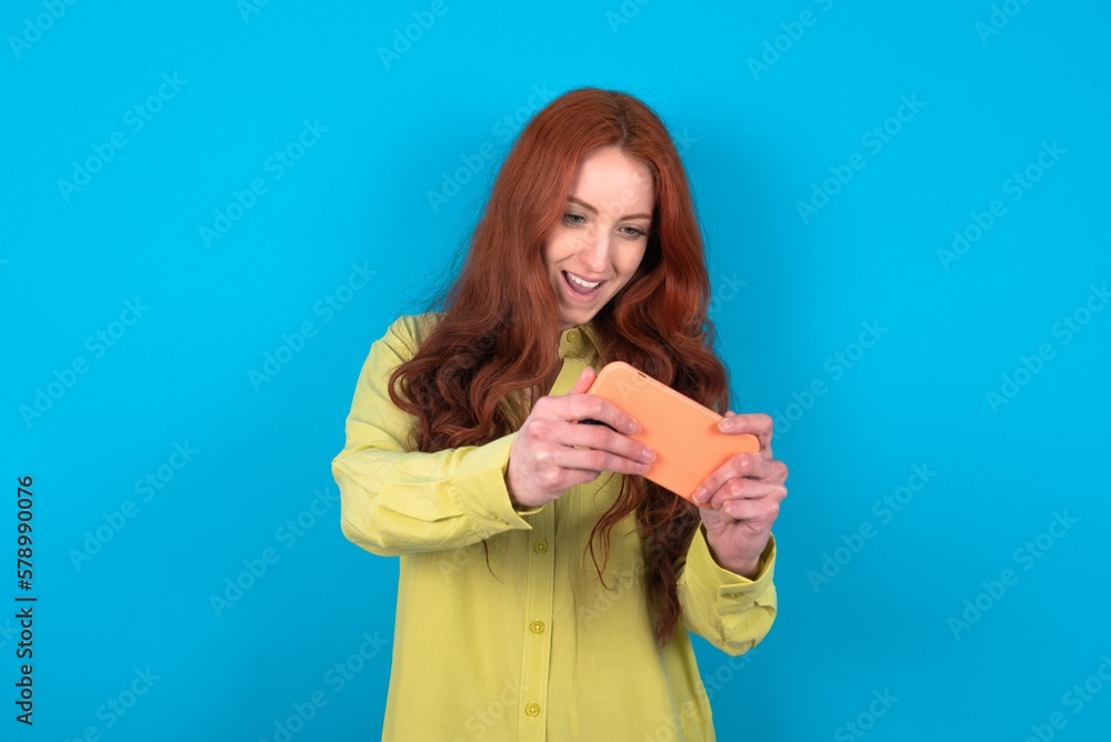 Portrait of an excited young woman wearing green sweater over blue background playing games on mobile phone. Portrait of an excited young woman wearing green sweater over blue background playing games on mobile phone.