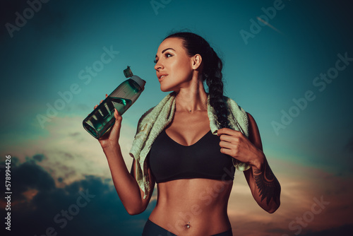 Young sports woman standing with bottle of water after training, tattoo on body