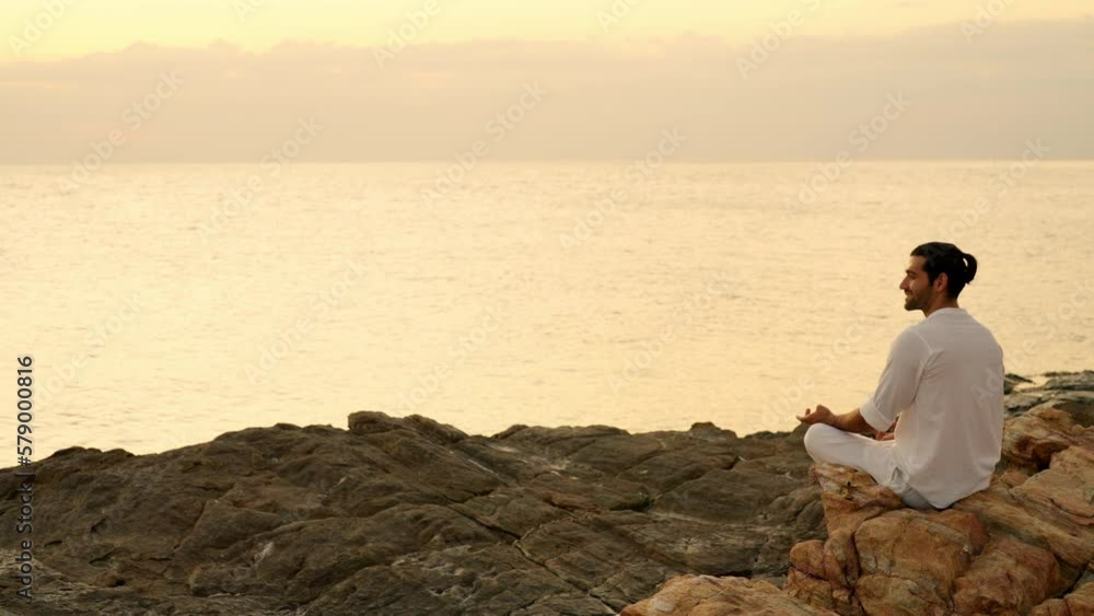 Caucasian man practicing meditation prayer with ocean nature on rocky coastal hill at summer sunset. Wellness people do outdoor self relaxing yoga exercise. Mental health care and motivation concept. Caucasian man practicing meditation prayer with ocean nature on rocky coastal hill at summer sunset. Wellness people do outdoor self relaxing yoga exercise. Mental health care and motivation concept.