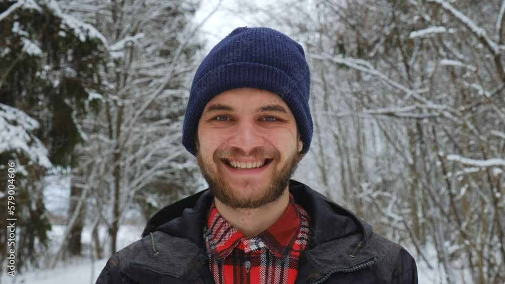 Young handsome Caucasian man stands in snowy winter forest and smiles. European guy in park during snowfall, close-up portrait. Happy person wears dark blue knitted hat, jacket and red plaid shirt. Young handsome Caucasian man stands in snowy winter forest and smiles. European guy in park during snowfall, close-up portrait. Happy person wears dark blue knitted hat, jacket and red plaid shirt.