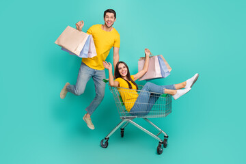 Photo of funny excited married couple wear yellow t-shirts riding shopping tray holding bargains isolated teal color background