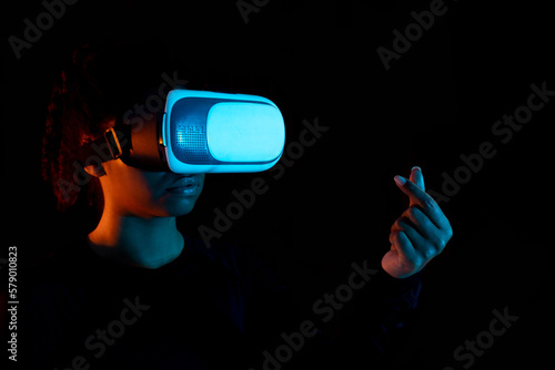Black woman wearing virtual reality goggles and making heart sign with hand in black light, studio shot