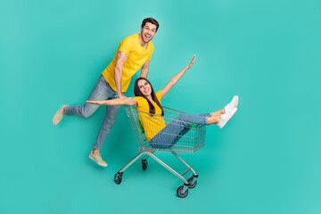 Photo of pretty carefree married couple wear yellow t-shirts riding shopping tray having fun isolated teal color background