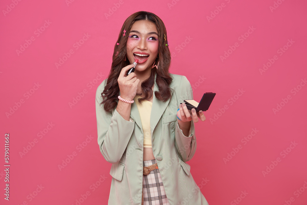 Naklejka premium Makeup lipstick woman ready looking in pocket mirror. beautiful trendy young asian female fashion model putting makeup on pink background.