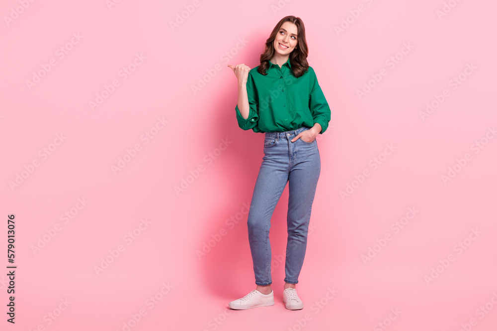 Full size photo of friendly cheerful girl wear green shirt arm in pocket look indicating at sale empty space isolated on pink background Full size photo of friendly cheerful girl wear green shirt arm in pocket look indicating at sale empty space isolated on pink background