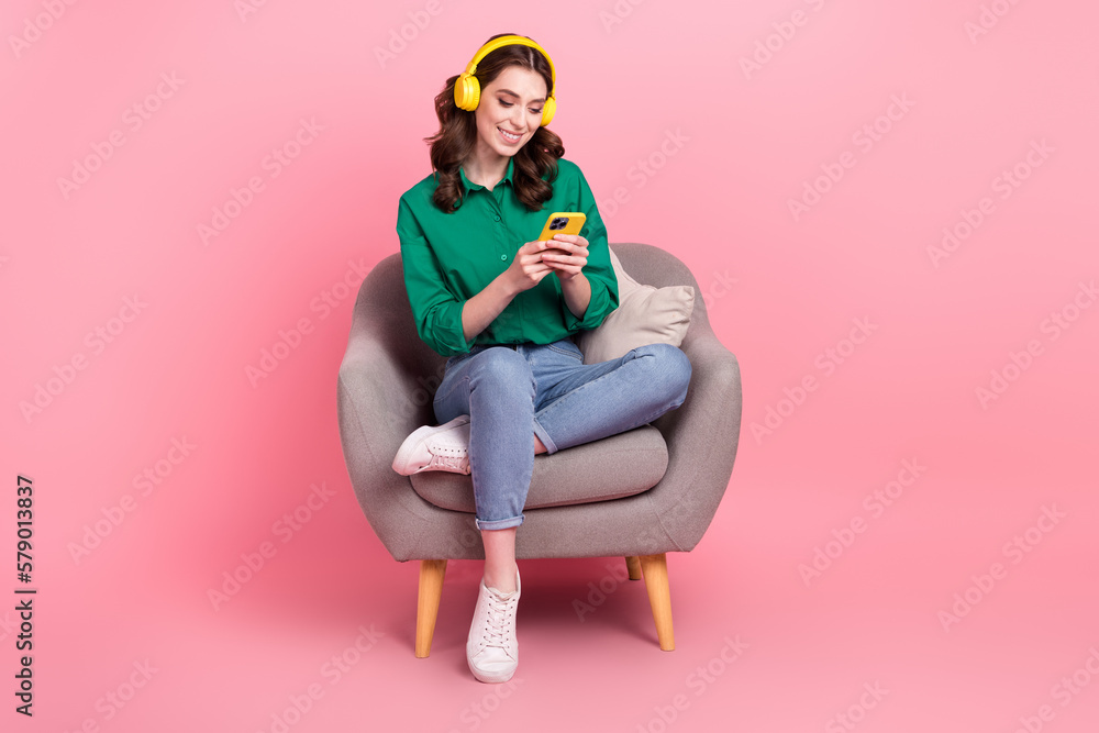 Full length photo of sweet shiny woman wear green shirt sitting couch enjoying songs modern gadget isolated pink color background
