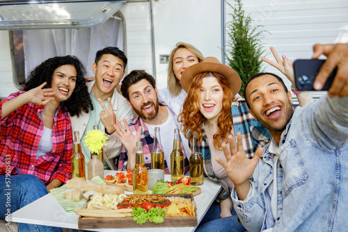 Mixed race friends taking selfie on mobile phone, outdoor party on travel trailer background. Rest outside the city. Happy people enjoy. Weekends in the summer time.
