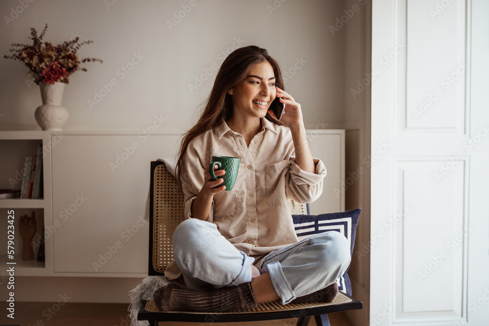 Smiling pretty young asian woman calls by smartphone, speaks, sits in armchair, enjoys cup of drink