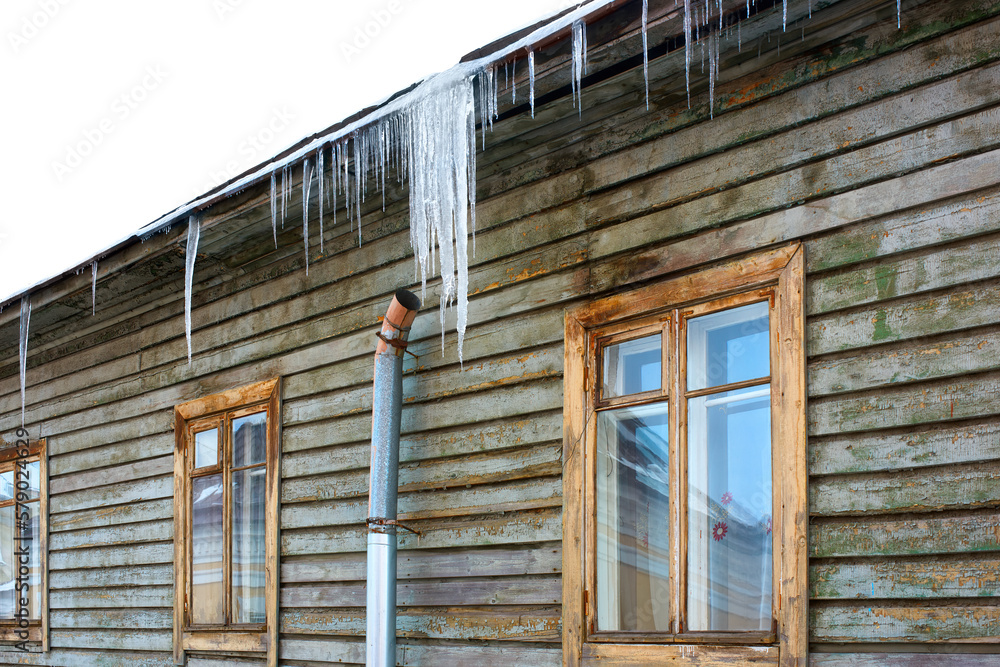 Naklejka premium Windows of an old wooden house with icicles on the roof. Peeling paint on the wall of a wooden house. Large, dangerous icicles on the roof of a building.