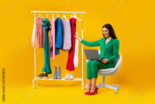 Online shopping. Stylish young lady using laptop and purchasing clothes online, sitting near clothing rail