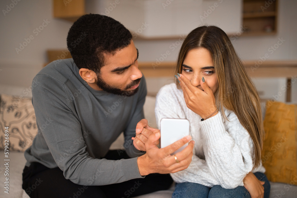 Sad angry millennial arab guy shows smartphone to european woman in interior room, close up Sad angry millennial arab guy shows smartphone to european woman in interior room, close up