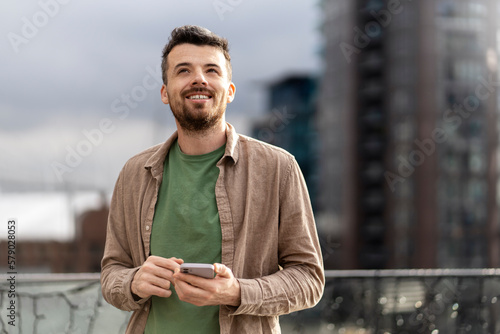 Handsome smiling middle aged hispanic man holding smartphone using mobile app shopping online, ordering food standing on the street. Modern hipster guy text messaging. Mobile banking concept 