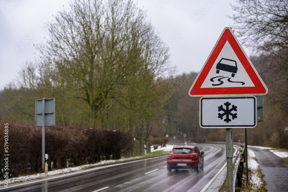 Car passing on a wet road in winter with a slippery road and ice warning sign Car passing on a wet road in winter with a slippery road and ice warning sign