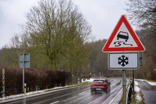 Wall Mural Car passing on a wet road in winter with a slippery road and ice warning sign