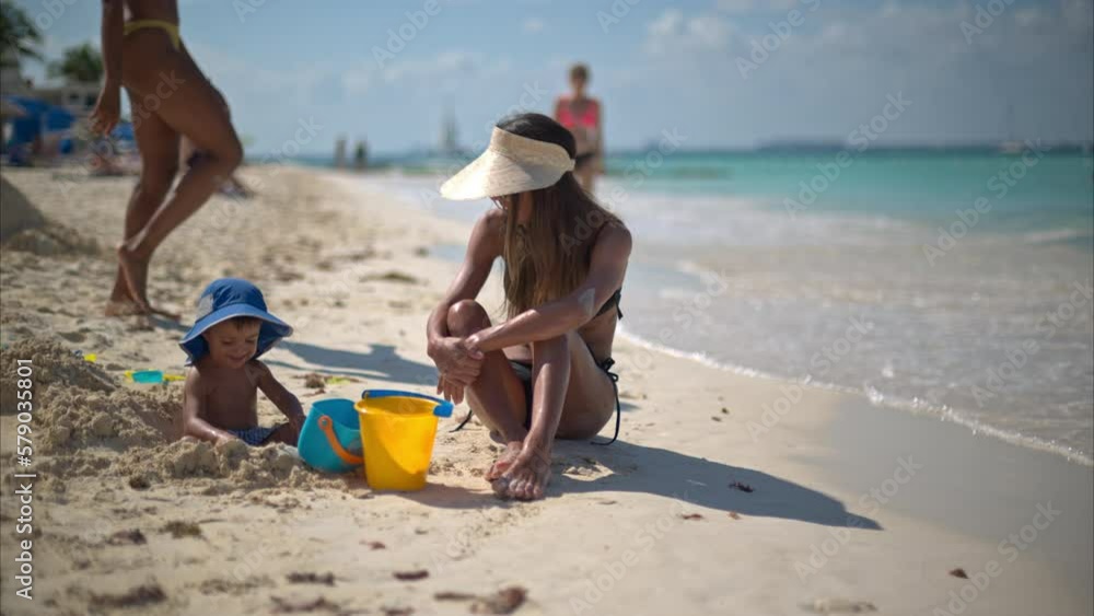 Cute hispanic toddler playing on the beach with the sand under his mother's watch while people walk by Cute hispanic toddler playing on the beach with the sand under his mother's watch while people walk by
