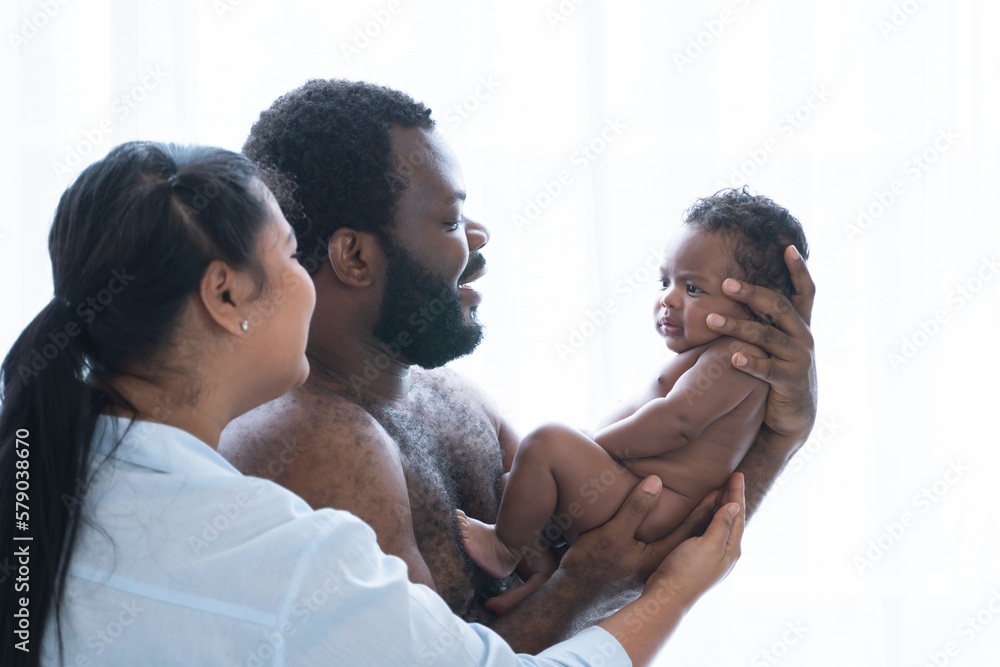 Fototapeta premium African Nigerian father and Asian mother carrying holding cute newborn baby in parents hands, smiling looking at infant with love at home, standing. Multiracial family bonding and child care concept