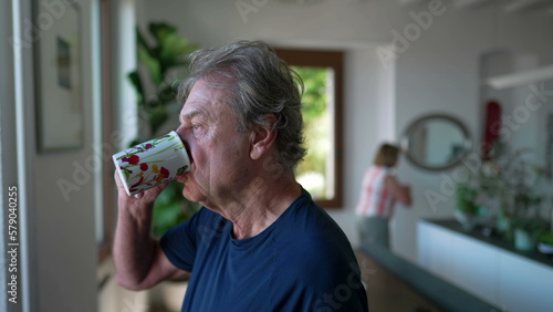 One mature older person drinks coffee or tea standing by window looking out in contemplation. 70s senior man with pensive expression