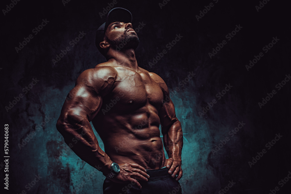 Fototapeta premium Young strong man bodybuilder in cap on wall background, dramatic colors