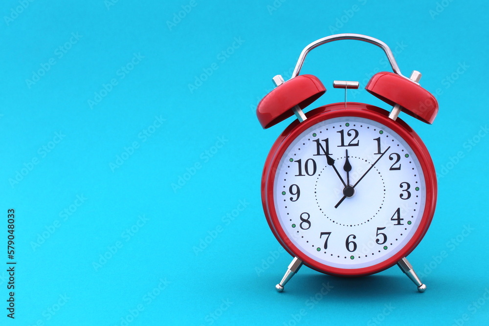 A red alarm clock stands on a blue background. The time shows five minutes to twelve A red alarm clock stands on a blue background. The time shows five minutes to twelve