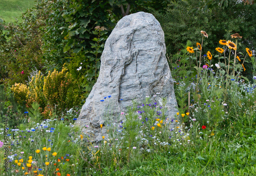 Ein kleiner, von bunten Blumen umgebener Hinkelstein vor einem Busch auf einer Wiese photo