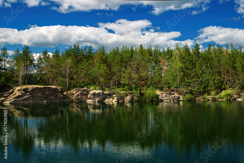 Fototapeta Naklejka Na Ścianę i Meble -  A forest lake with rocky shores and coniferous trees, with a blue sky and white clouds reflected in the water