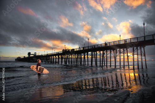 Young woman with a surfboard standing by the pier at sunset, Oceanside, California, USA photo