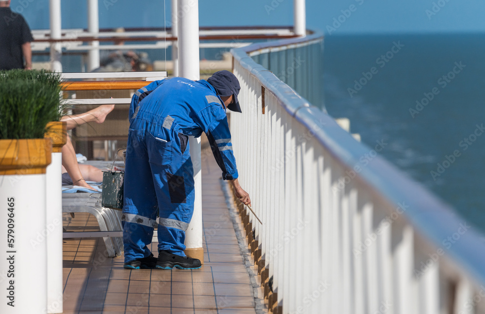 Guests relax as crew member paints the railing around deck of cruise ship as it sails at sea