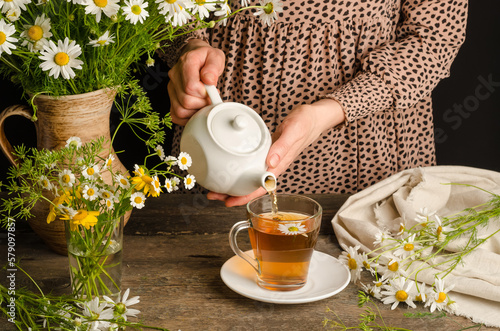 Woman pouring chamomile herbal tea on dark wooden background