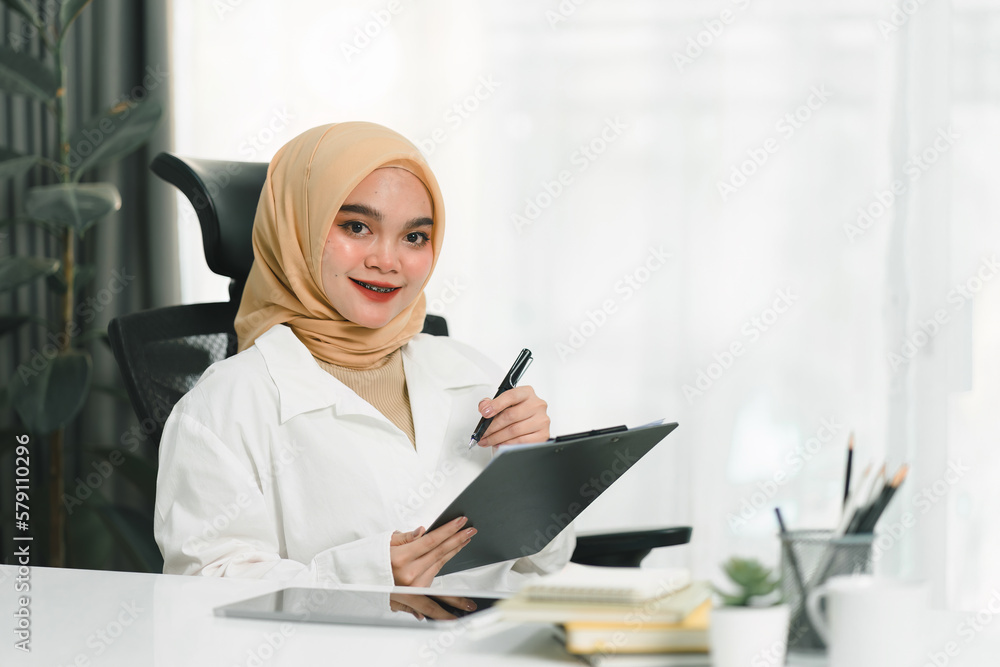A smiling and confident young Muslim businesswoman auditor is writing on a clipboard and signing a contract document at her desk in a modern office. A smiling and confident young Muslim businesswoman auditor is writing on a clipboard and signing a contract document at her desk in a modern office.