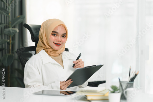 A smiling and confident young Muslim businesswoman auditor is writing on a clipboard and signing a contract document at her desk in a modern office.