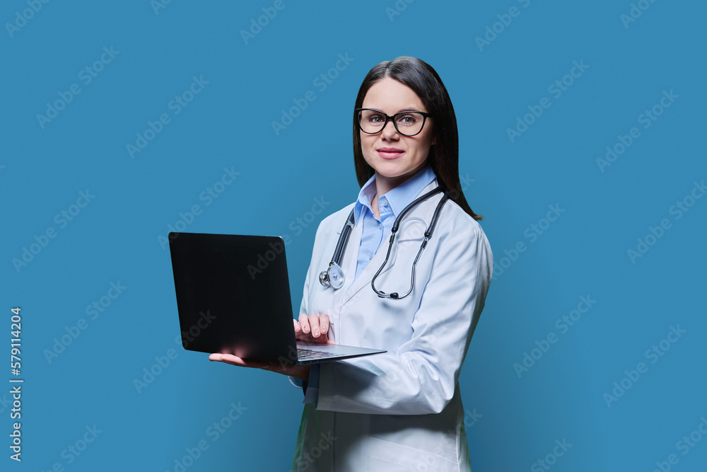 Young friendly female doctor using laptop on blue background Young friendly female doctor using laptop on blue background