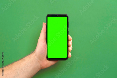 Mobile phone in young man hand. Use smartphone with blank green screen on green color studio background. Male showing black phone with chroma key display. Close up. Mock up of smart phone for Ad, app