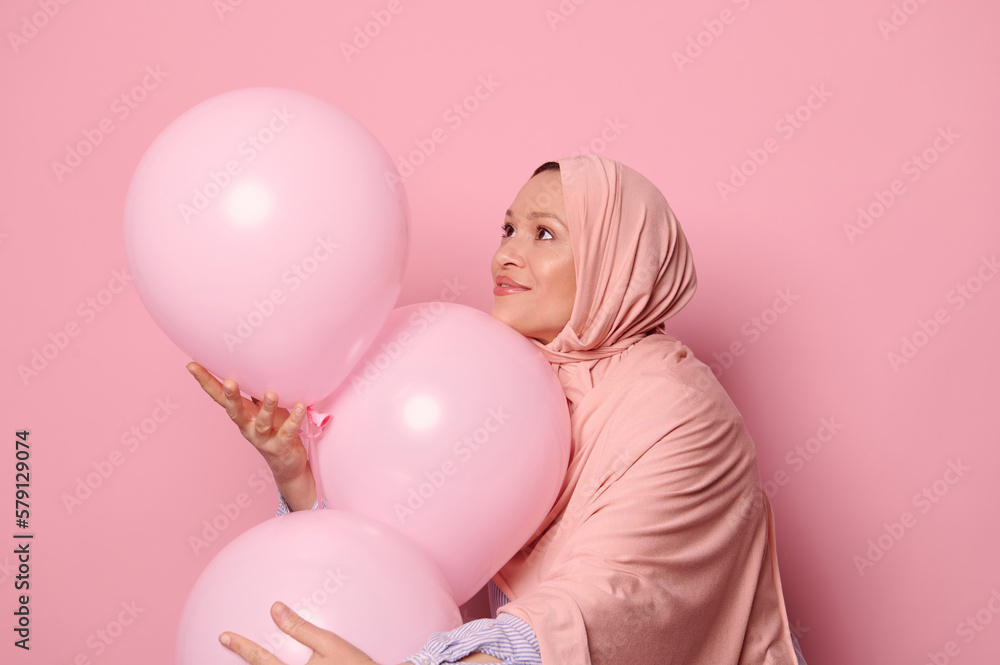 Happy Middle-Eastern Muslim woman in a pink hijab, catching pink pastel inflatable helium party balloons, isolated over pink color background.