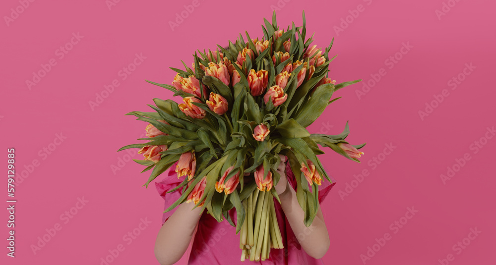 Cute smiling child holding a beautiful bouquet of tulips in front of his face isolated on pink. Little toddler girl gives a bouquet to mom