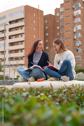 Portrait of two young woman students in university during break, carrying laptop and books in hand, outdoor education concept.