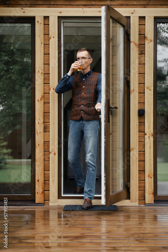 Middle age man comes out on the terrace of a small wooden house with a glass in hand