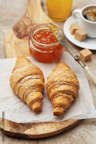 Freshly baked croissants on wooden cutting board, jam, cup of coffee and orange juice over beige concrete background. Healthy delicious breakfast. Close up view