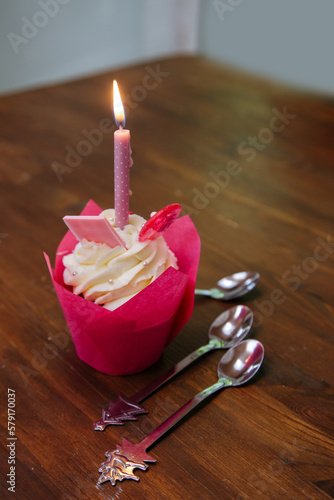 Tasty cupcake for birthday party with candel and spoons on a brown wooden table. Cakes decorated with white cheese cream, pink lollipop and chocolate. For a girls party.