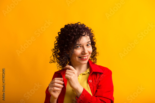Studio shot of positive and smiling pretty female. Curly girl touching her hair. Young woman in bright red shirt. Isolated on yellow background. Close up.
