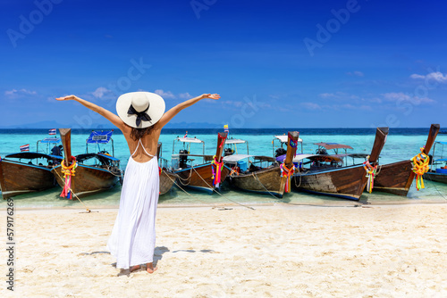 A happy tourist woman in a white dress stands in front of longtail boats on a paradise beach in Thailand during her tropical vacations photo