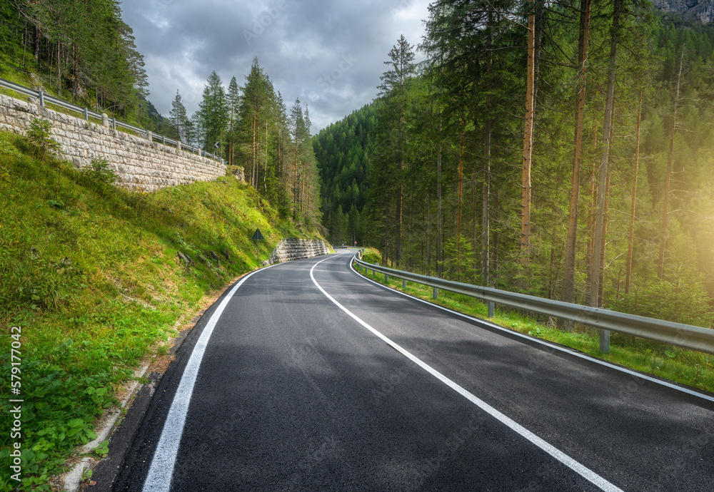 Road in green forest at sunset in summer. Dolomites, Italy. Beautiful mountain roadway, tress, grass, high rocks, blue sky with clouds. Landscape with empty highway through the wood in spring. Travel - obrazy, fototapety, plakaty 
