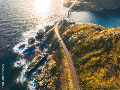 Aerial view of bridge, car, sea with waves and mountains at sunset in Lofoten Islands, Norway. Landscape with beautiful road, water, rocks and stones, golden sunlight. Top view from drone of highway